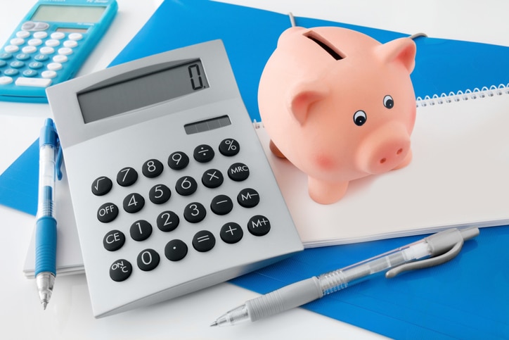A piggy bank, pens, and calculator on top of a notebook on a desk containing advice on saving money.